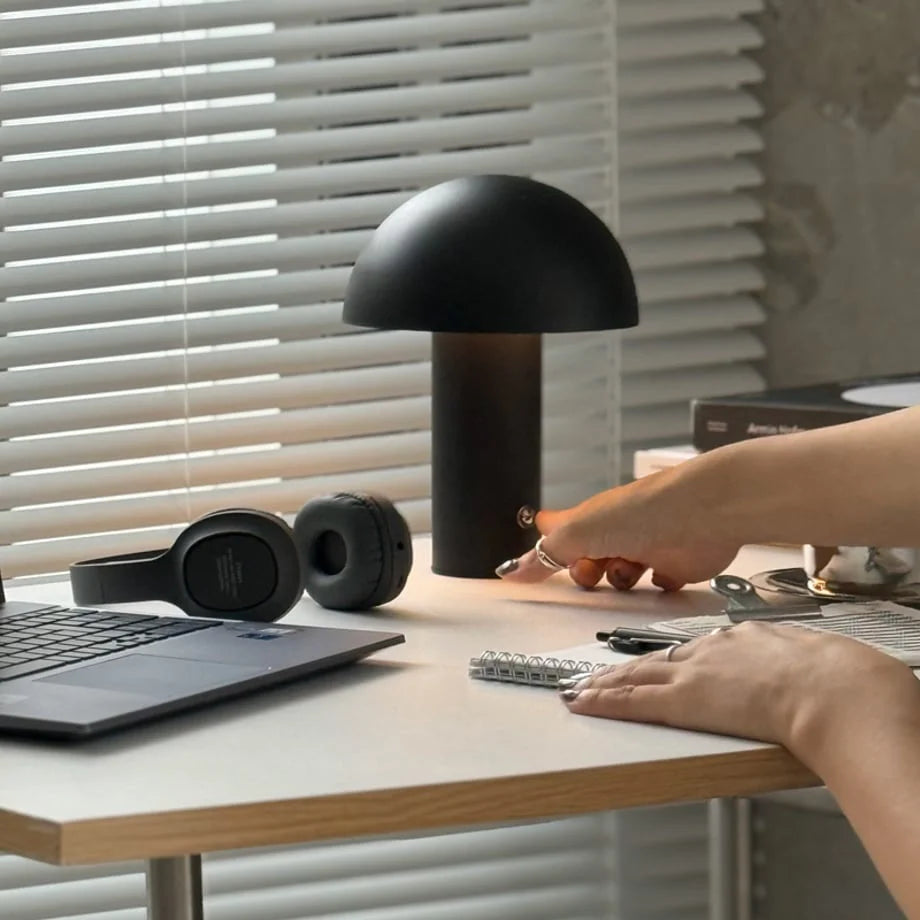 Close-up of a hand touching the base of the black mushroom lamp to adjust its brightness on a desk - RoomDen.