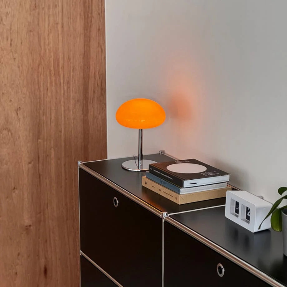 The orange mushroom table lamp glowing on a black console next to a stack of books and a flip clock - RoomDen.