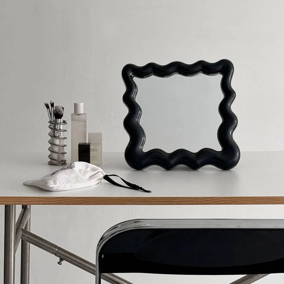 Black wavy frame mirror sitting on a white vanity desk with makeup brushes and bottles - RoomDen