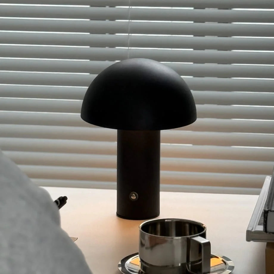The black minimalist mushroom lamp casting a focused glow on a desk with a stainless steel cup - RoomDen.