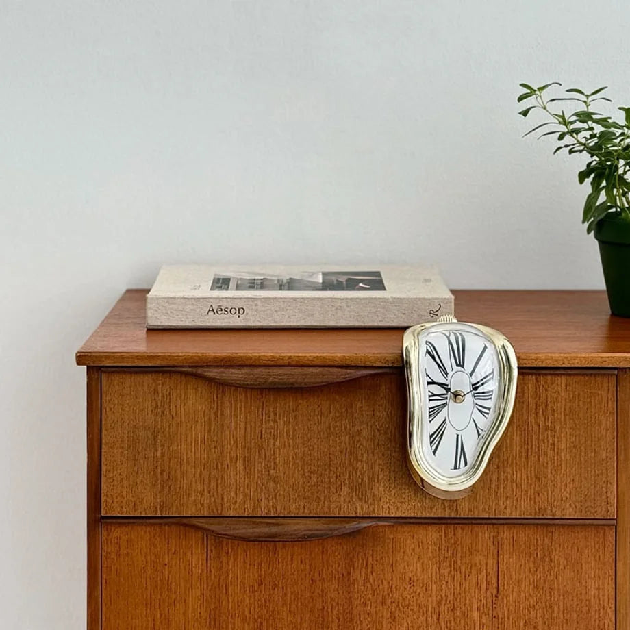 Gold Surrealist Melting Clock placed on the edge of a vintage wooden dresser. - RoomDen