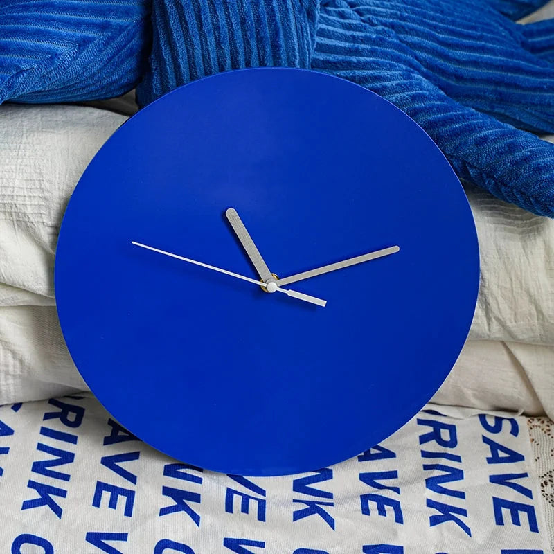 Close-up of the Klein blue minimalist wall clock resting on a bed with blue and white textured pillows, showing its smooth matte finish - RoomDen.