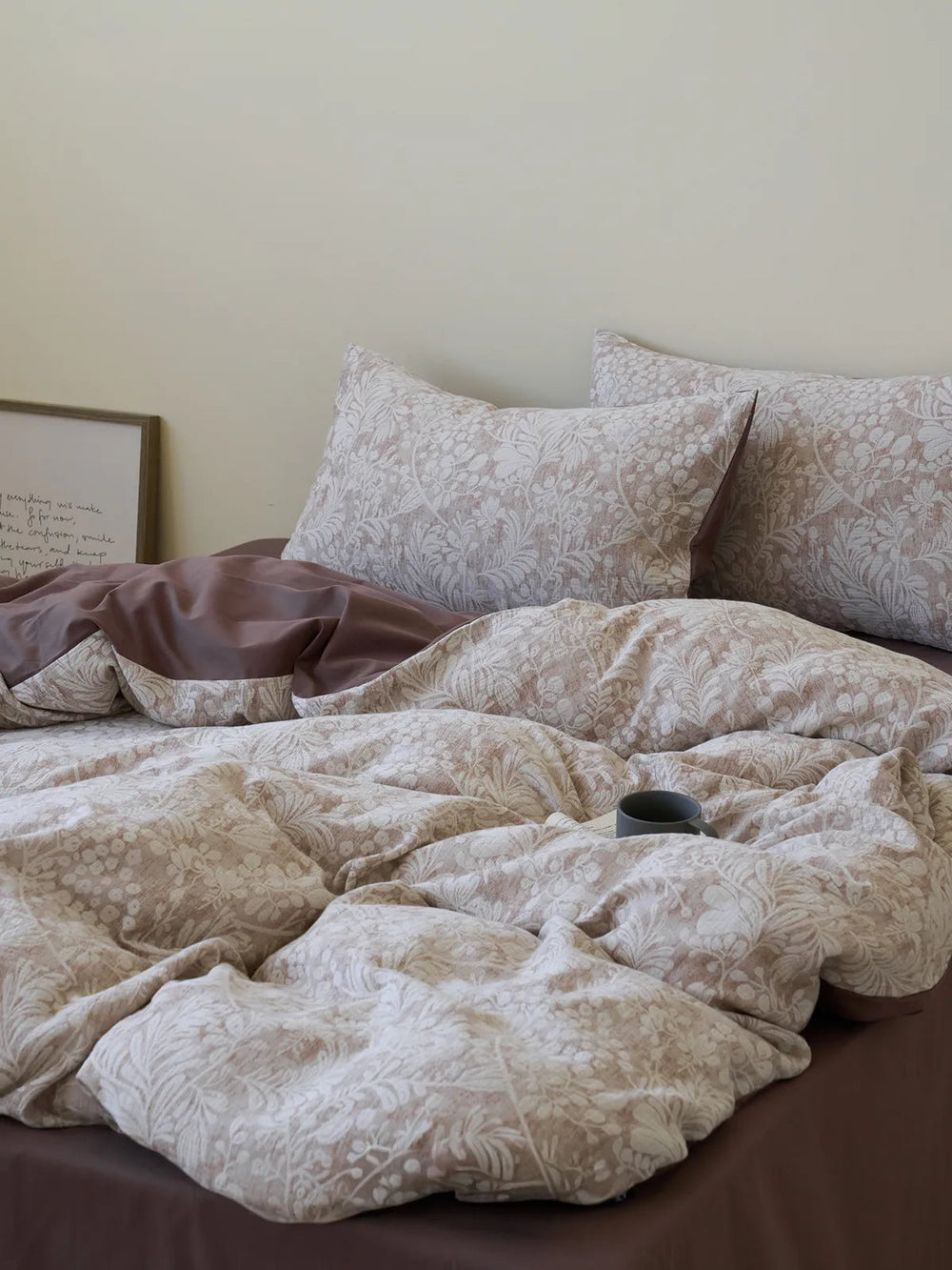 A cozy bed made with a Textured Vintage Botanical Washed Cotton Bedding Set, featuring a mocha brown flat sheet and a cream floral duvet cover. A grey mug rests on the bed. - RoomDen