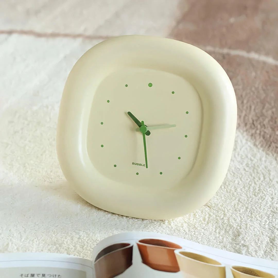 The Creamy Beige Puffy Bubble Table Clock sitting on a textured white surface next to an open magazine - RoomDen