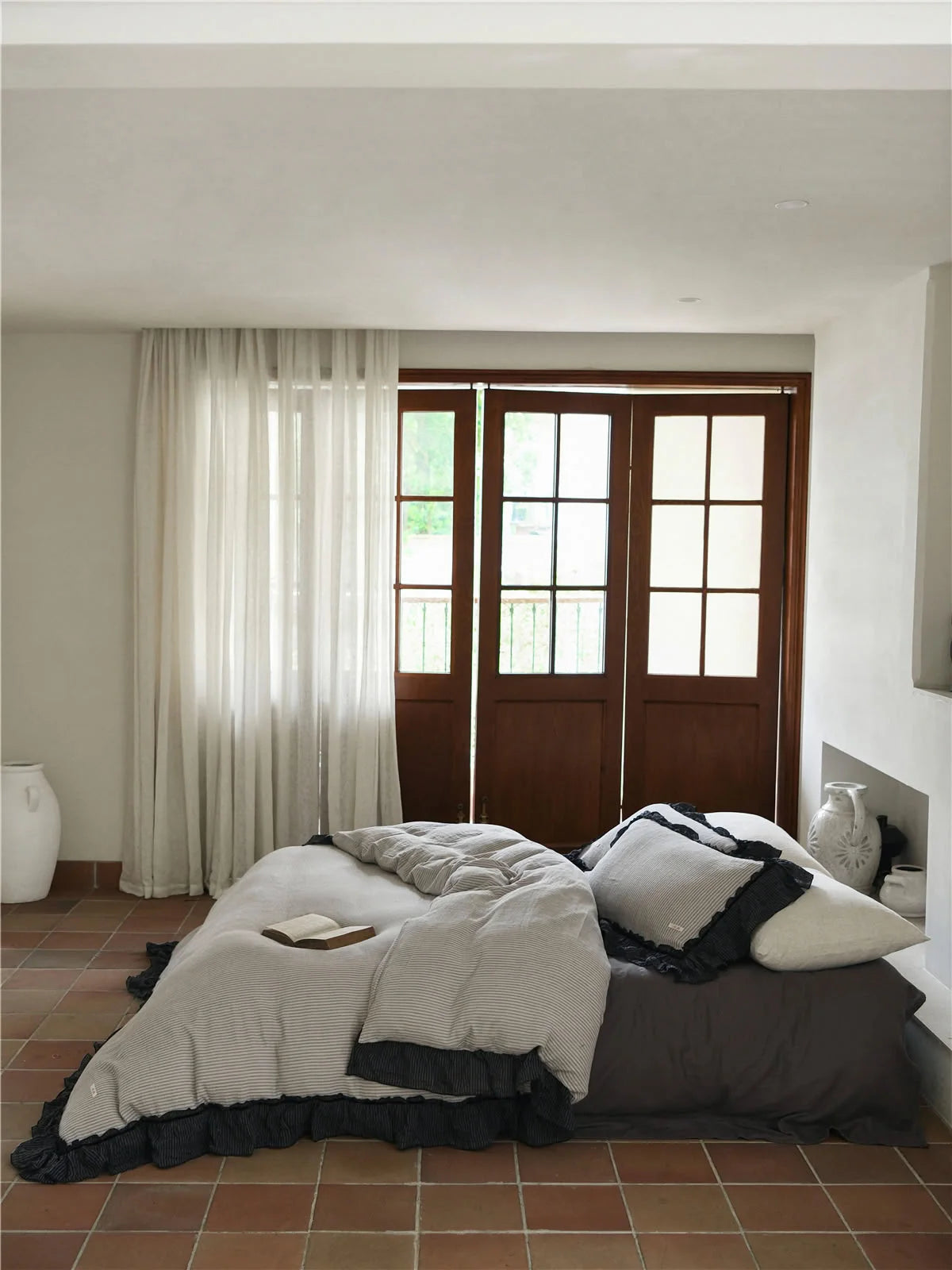 Full view of a bed with a grey and black pinstripe ruffle bedding set in a rustic room with terracotta floors. - RoomDen