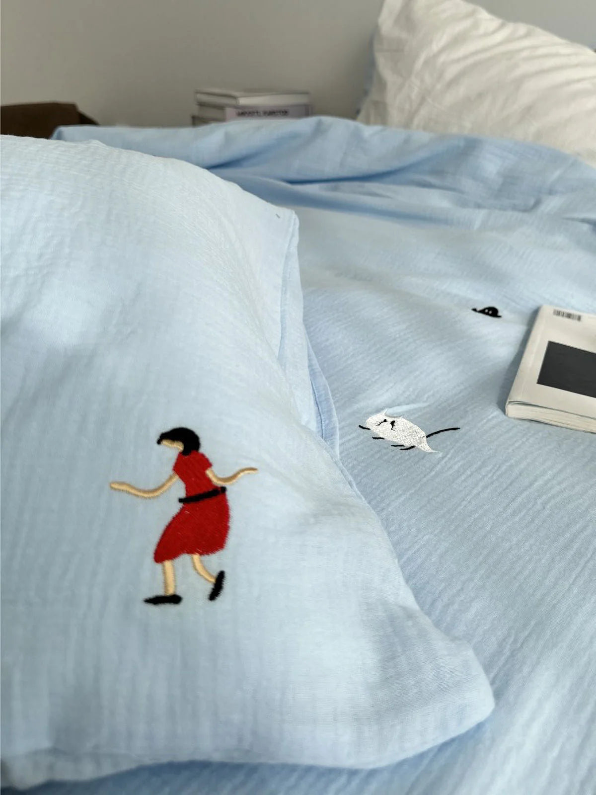Close-up shot on the pillowcase of the sky blue bedding, showing the detailed embroidery of a girl in a red dress.
