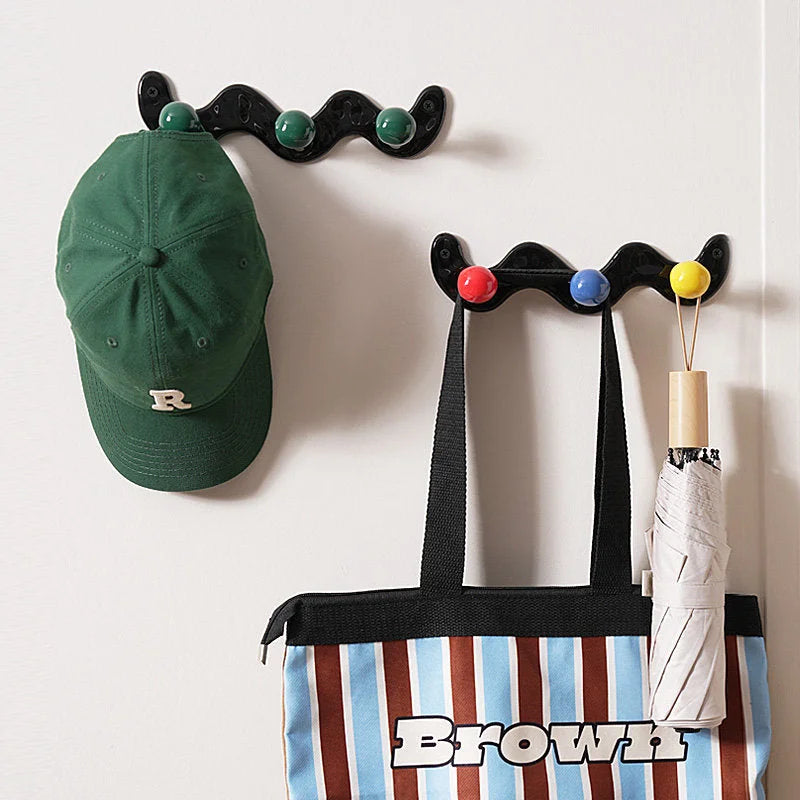 A green cap and a tote bag hanging from black wavy ceramic hooks with green and multicolor balls on a white door.