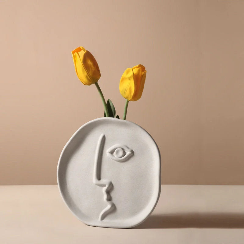 The round, plate-like abstract face vase holding two yellow tulips against a beige background - RoomDen.