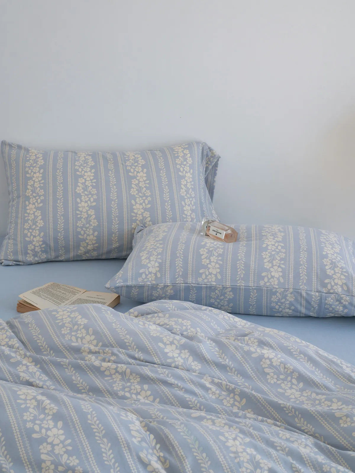 Close-up of the cornflower blue floral stripe pillowcases and duvet cover, with an open book and perfume bottle. - RoomDen