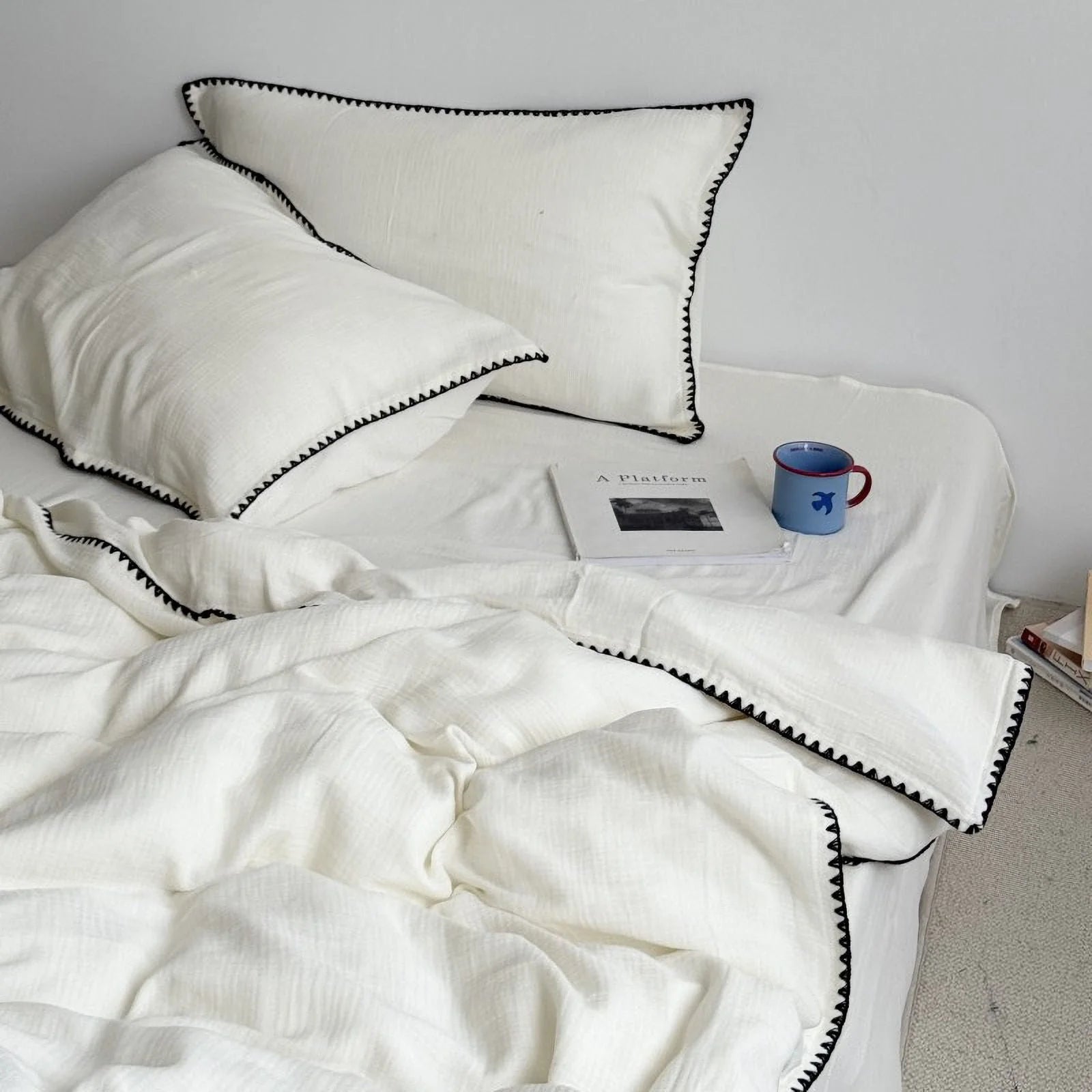 Cream white washed cotton bedding set with black embroidered edge, styled on a bed with a book and mug. - RoomDen