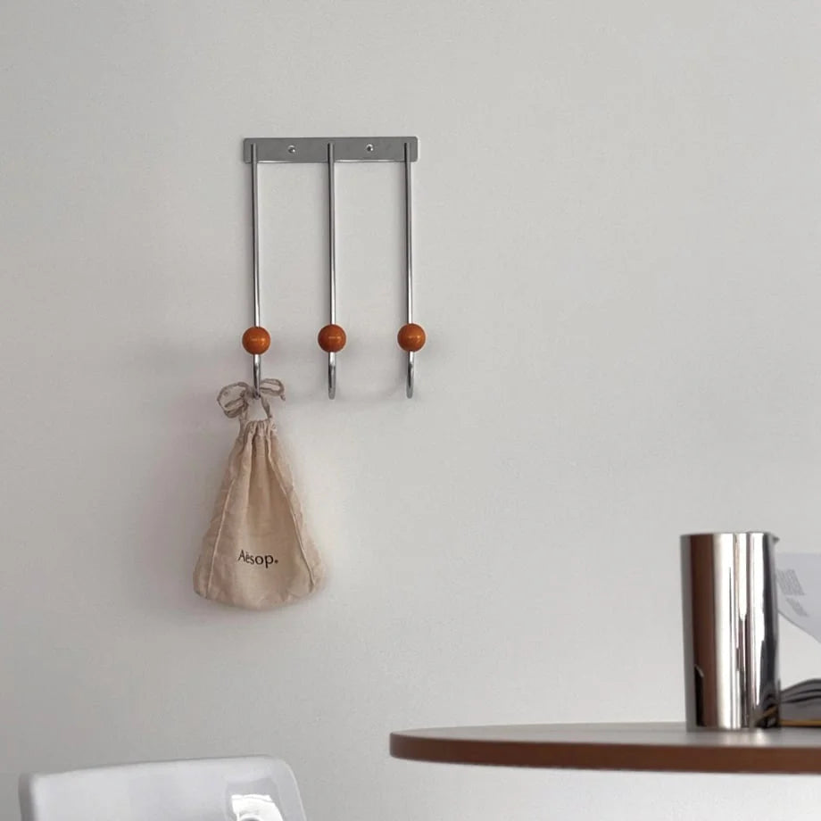 The Silver & Orange Metal Wall Hooks holding a beige Aesop pouch in a clean, white room setting. - RoomDen