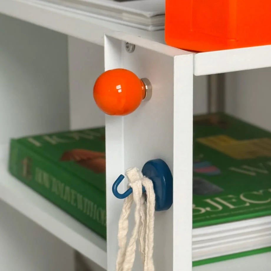 An orange magnetic hook and a blue hook with a loop attachment on a white shelf. - RoomDen