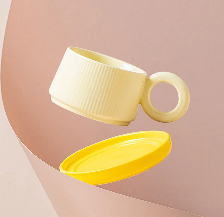 The Butter Yellow ribbed ceramic mug and its matching yellow saucer shown floating against a pink background. - RoomDen