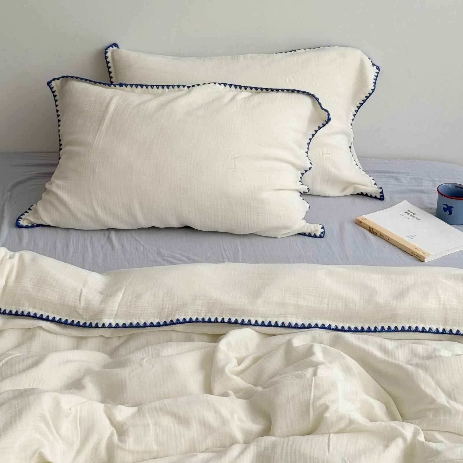 Full view of the cream white and blue embroidered washed cotton bedding set styled on a bed with a book. - RoomDen