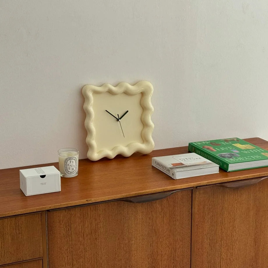 A cream-colored Wavy Squiggle Clock sitting on a wooden console table next to books and a candle. - RoomDen