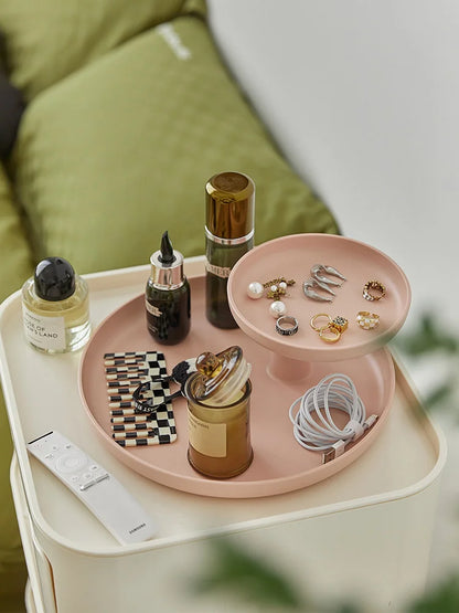 Blush pink two-tier organizer tray on a nightstand holding jewelry, cosmetics, and a charging cable. - RoomDen