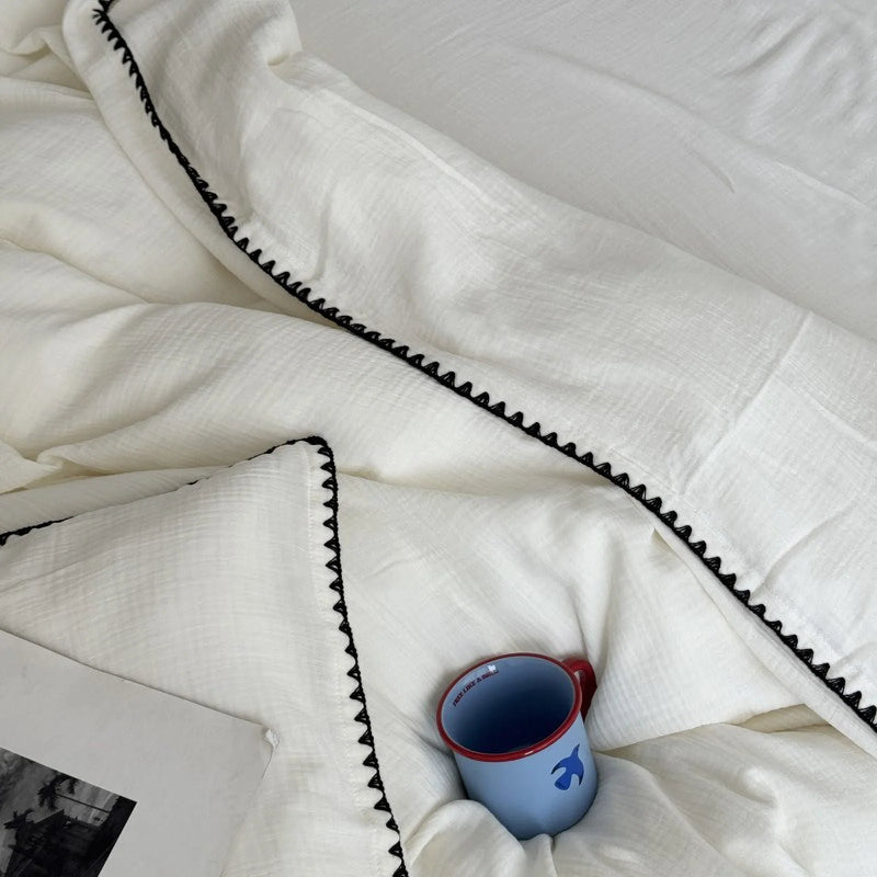 Overhead shot of a cream white washed cotton duvet with embroidered edge and a blue mug, light pooling softly across the surface in a 3-meter-squint test.