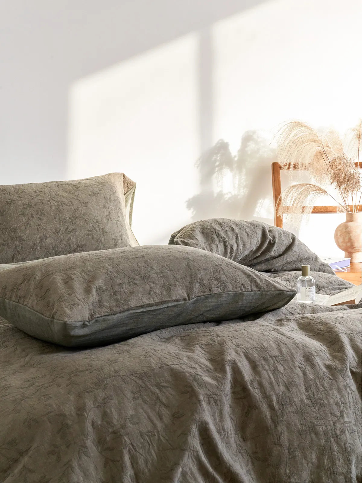 Angled view of a bed made with the earthy green botanical bedding, with sunlight casting shadows and a vase of pampas grass nearby.