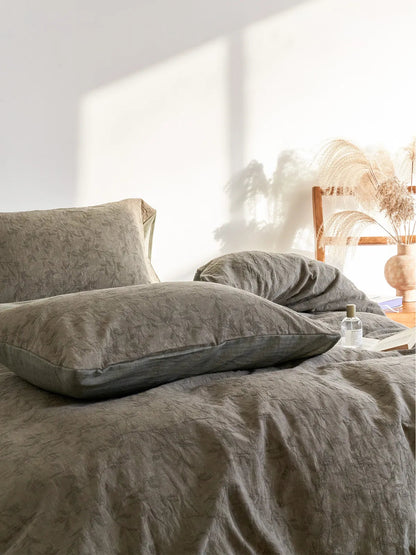 Angled view of a bed made with the earthy green botanical bedding, with sunlight casting shadows and a vase of pampas grass nearby.