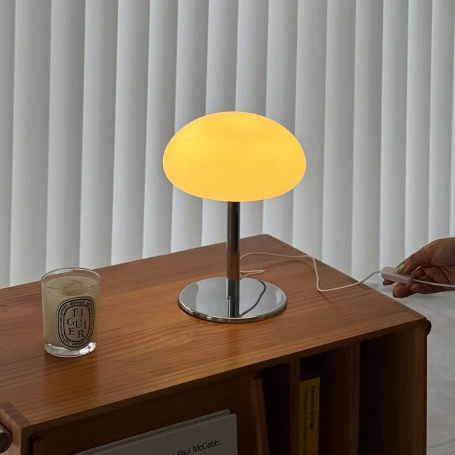 The white mushroom lamp emitting a warm yellow light, with a hand on the power switch - RoomDen.