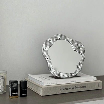 Molten Irregular Chrome Desktop Mirror placed on a stack of books next to beauty products - RoomDen