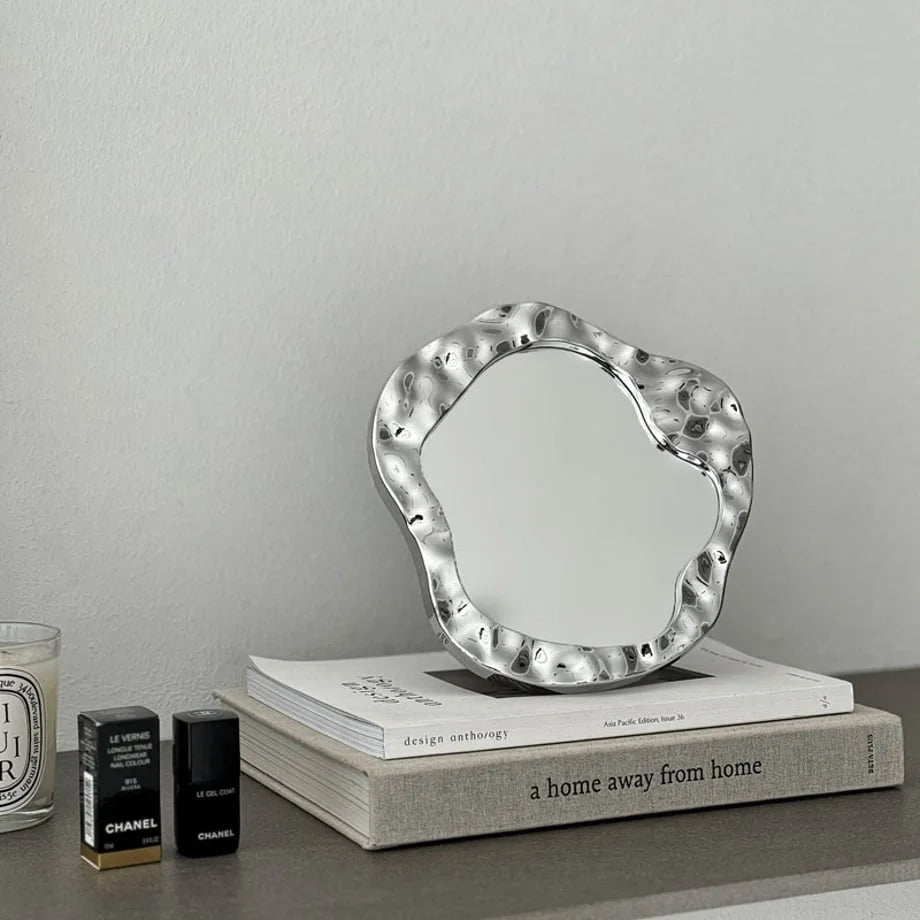 Molten Irregular Chrome Desktop Mirror placed on a stack of books next to beauty products - RoomDen