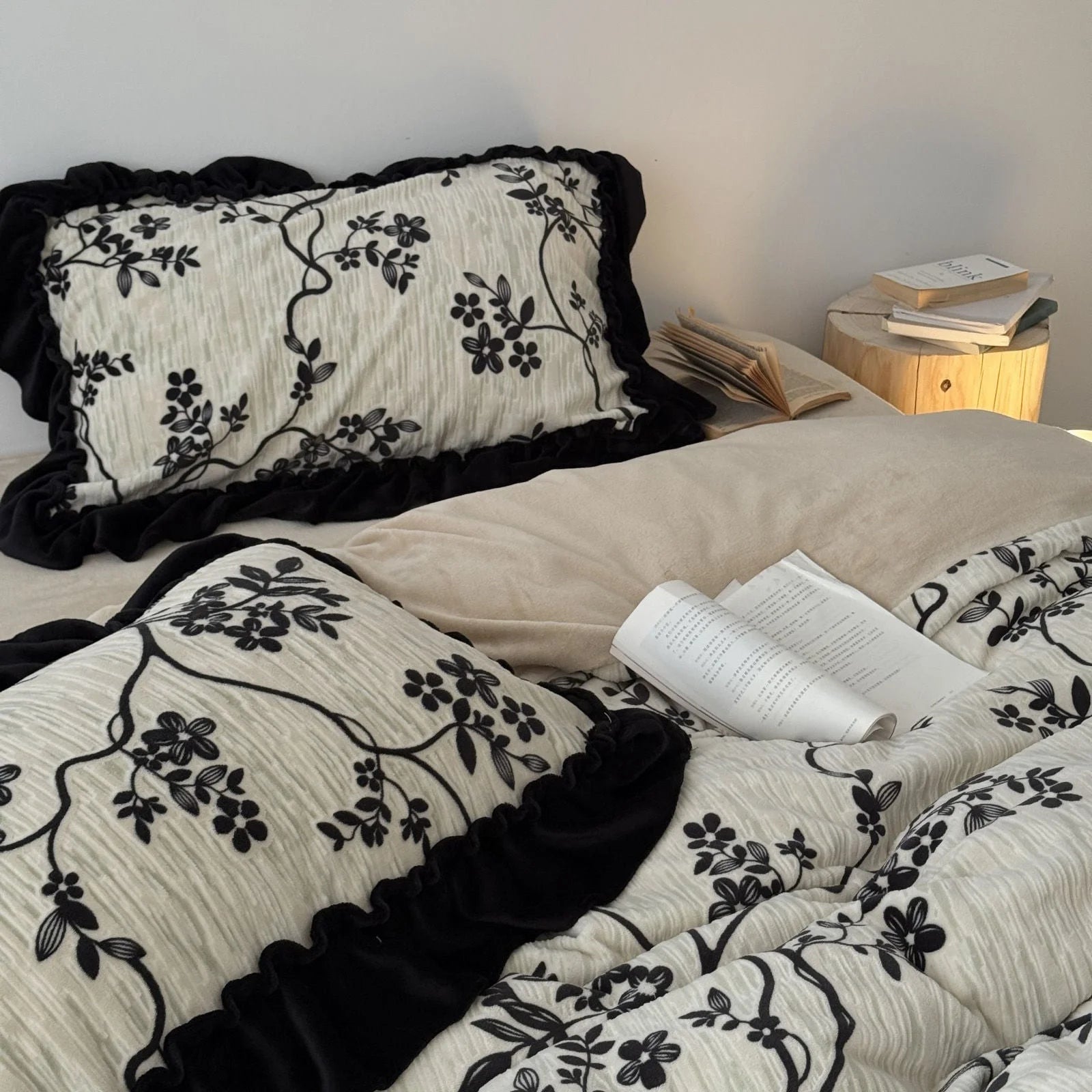 A styled shot of the cream and black floral bedding set, with stacked books and a lamp on a nightstand in the background - RoomDen.