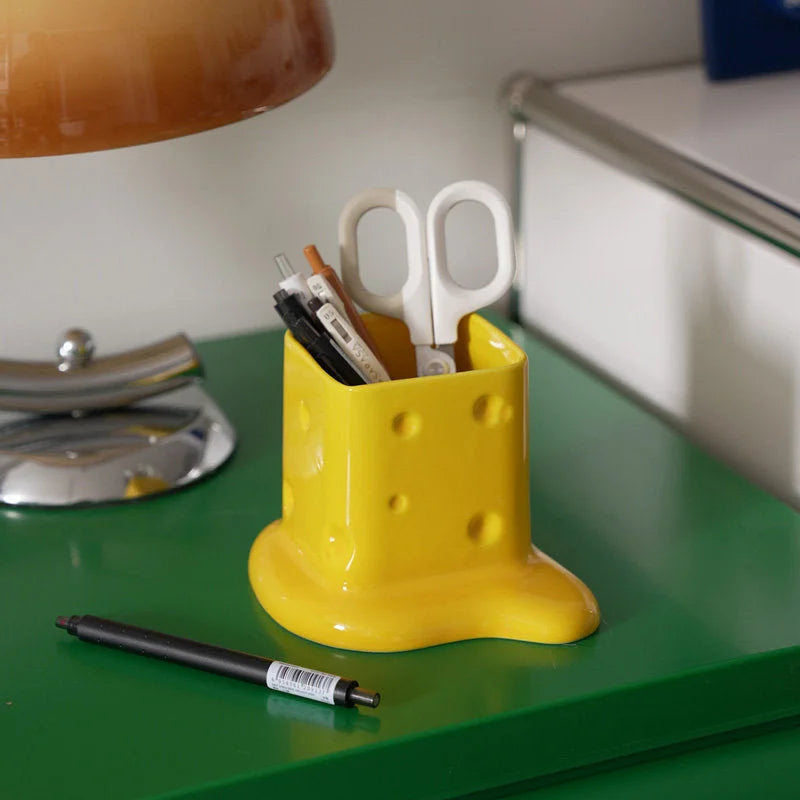 A yellow melting cheese ceramic organizer holding pens and scissors on a green desk next to a lamp. - RoomDen