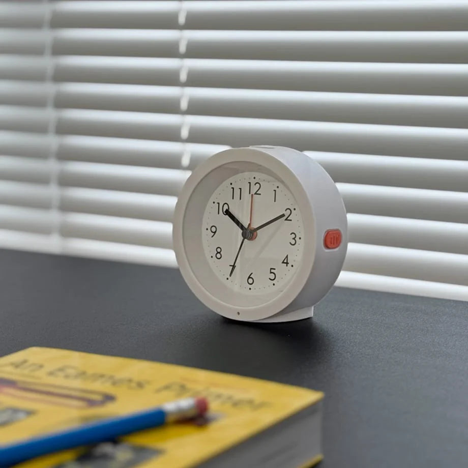 The white Minimalist Matte Analog Alarm Clock on a dark desk next to a book. - RoomDen