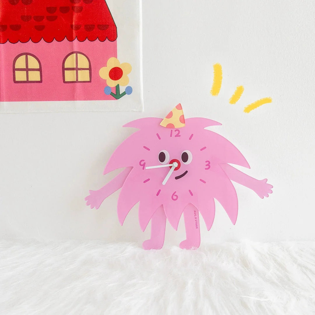 Playful pink monster-shaped clock standing on a white desk next to a colorful house illustration.