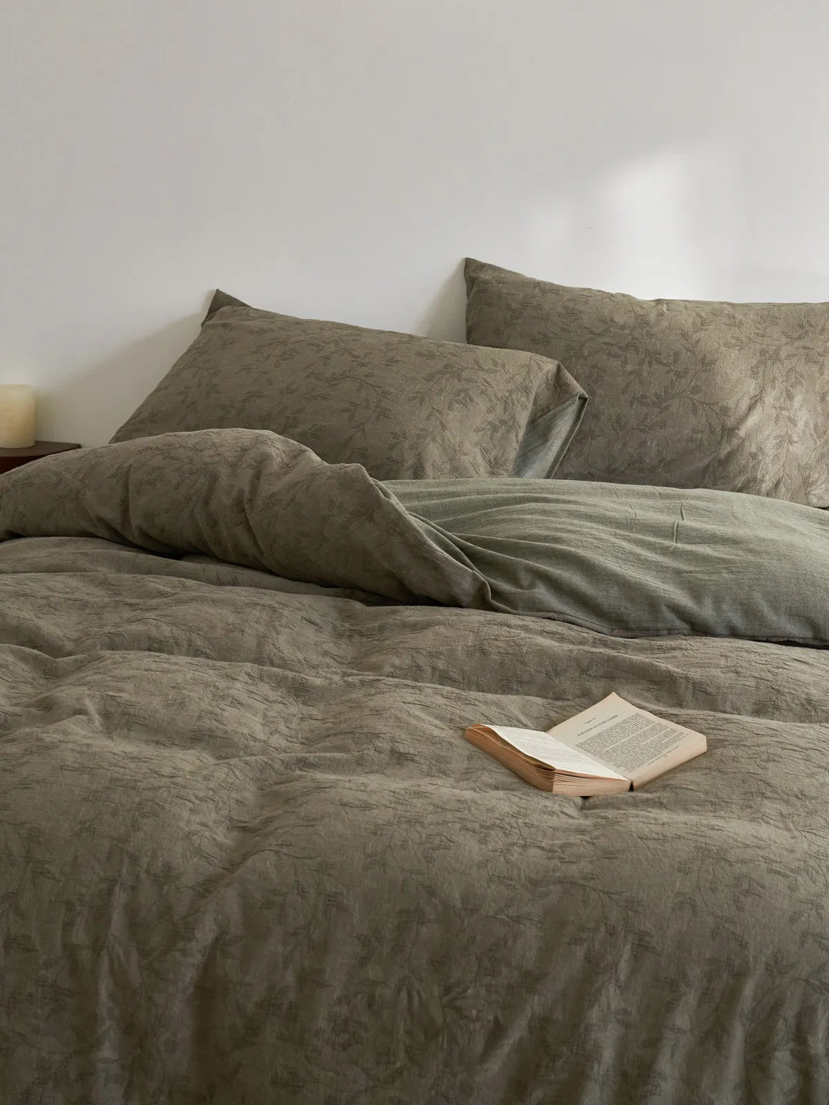 A cozy, rumpled bed with the earthy green botanical duvet cover and pillows, styled with an open book, creating a calm and inviting atmosphere.