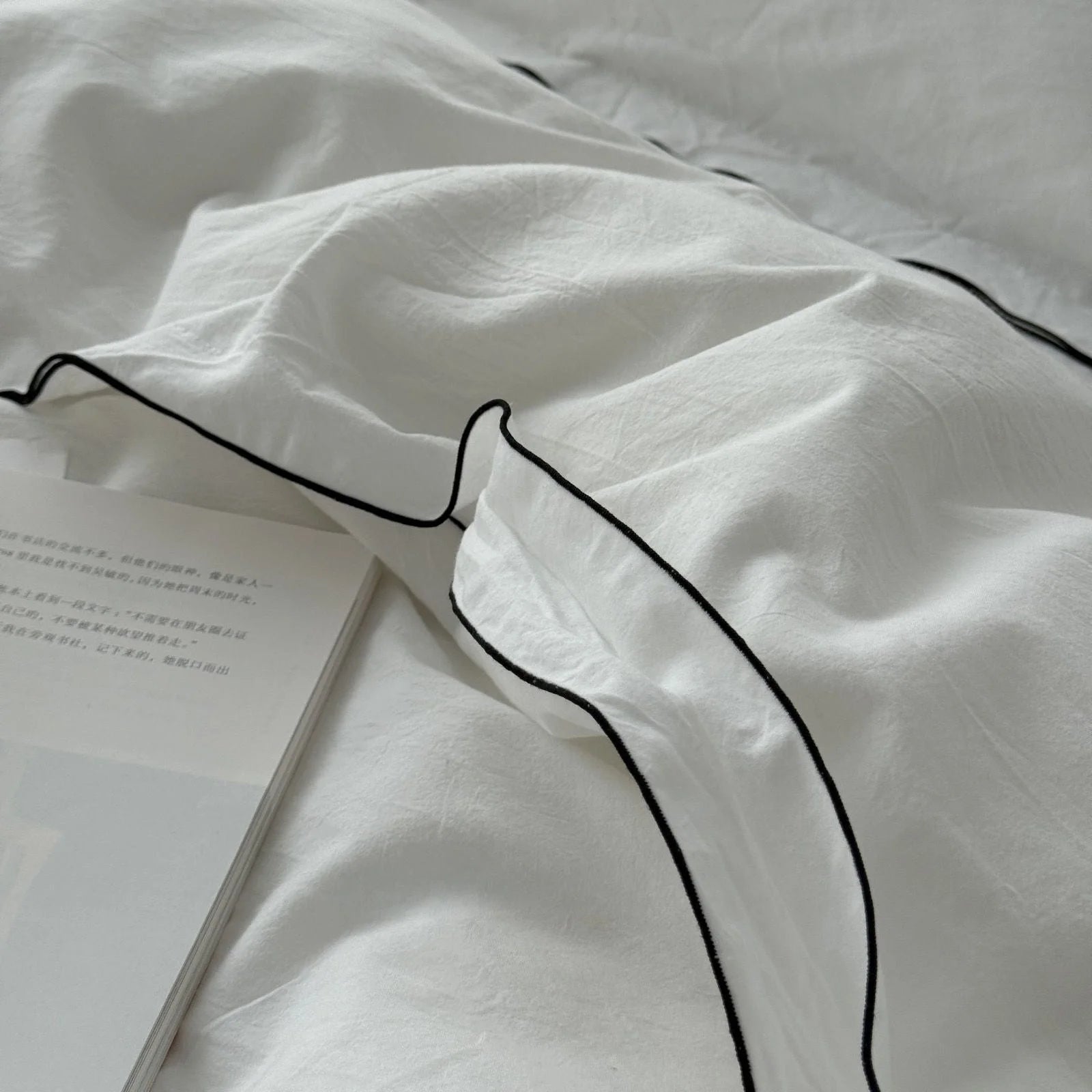 Extreme close-up on the corner of a white pillowcase, showing the fine black piping and the textured weave of the washed cotton. - RoomDen