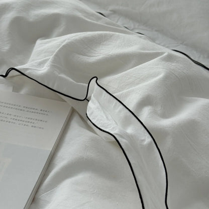Extreme close-up on the corner of a white pillowcase, showing the fine black piping and the textured weave of the washed cotton. - RoomDen