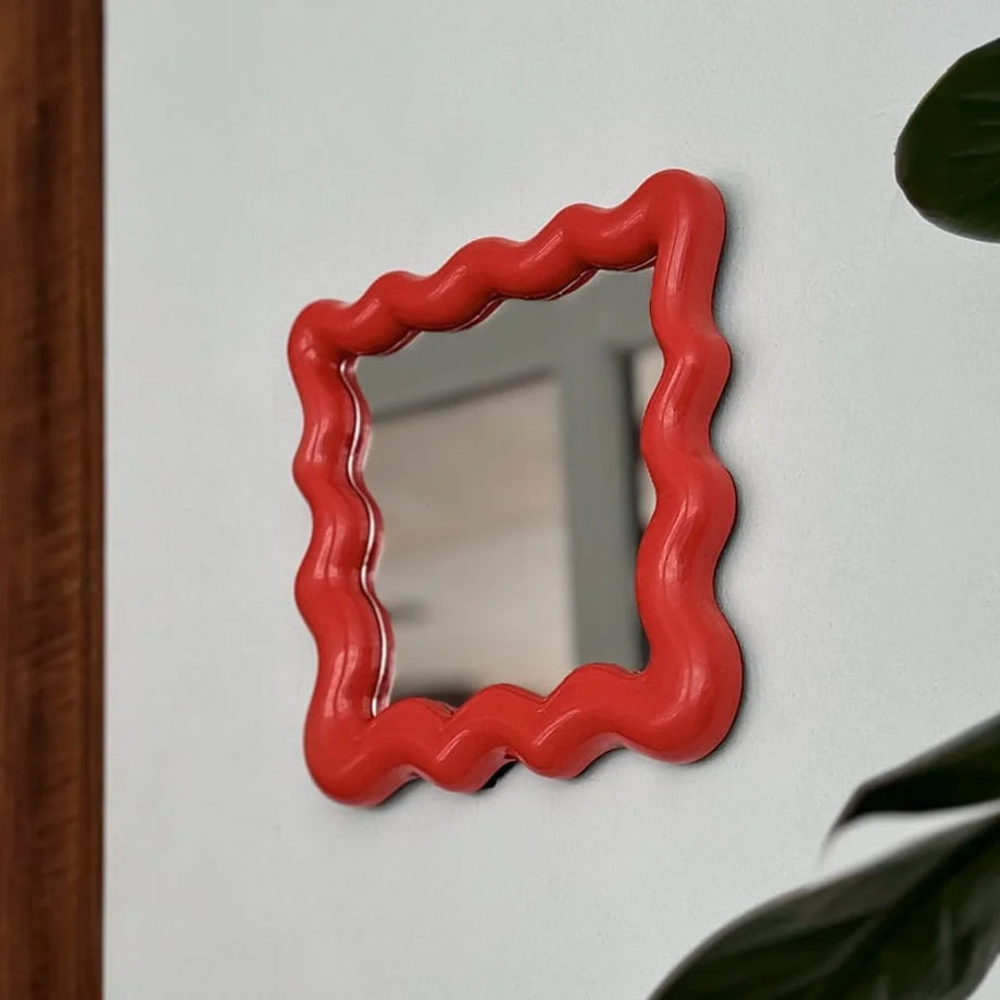 Angled shot of the red squiggle frame mirror on a wall, with a plant leaf in the foreground - RoomDen