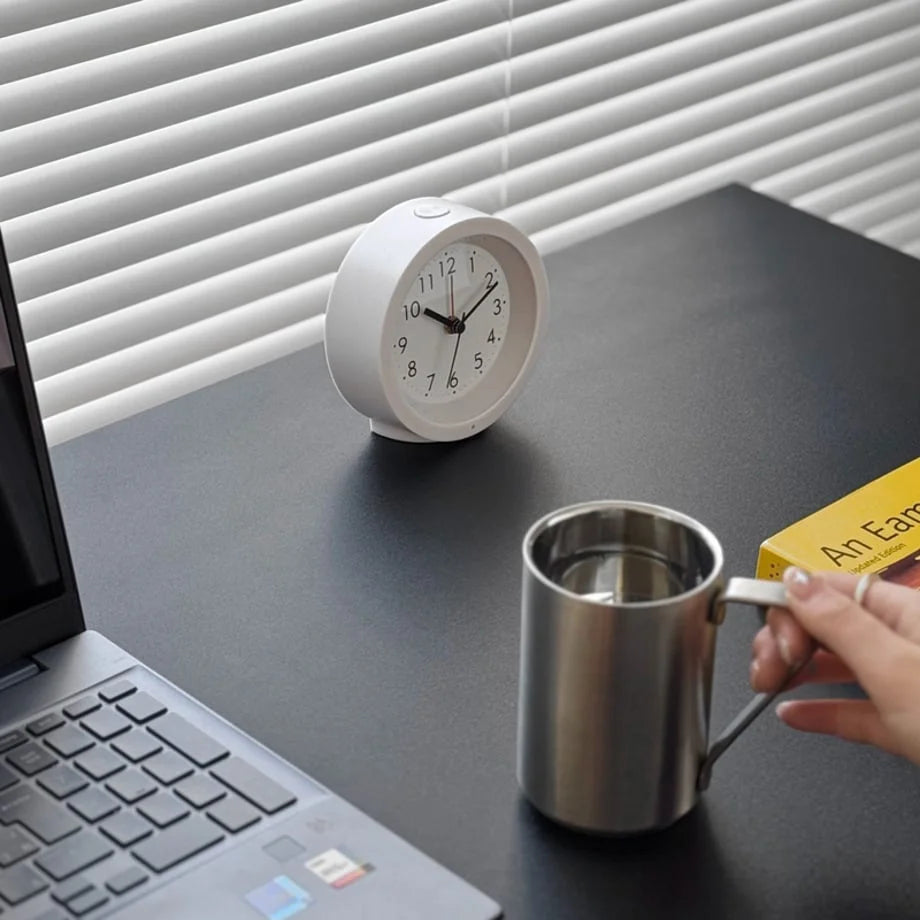 Lifestyle image of the white analog alarm clock on a desk with a person reaching for a mug. - RoomDen