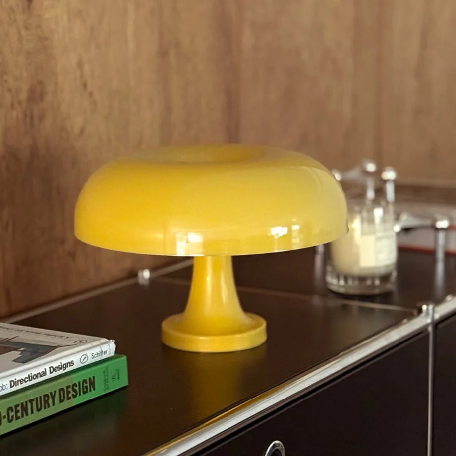 The marigold yellow mushroom lamp glowing warmly on a black surface next to design books - RoomDen.