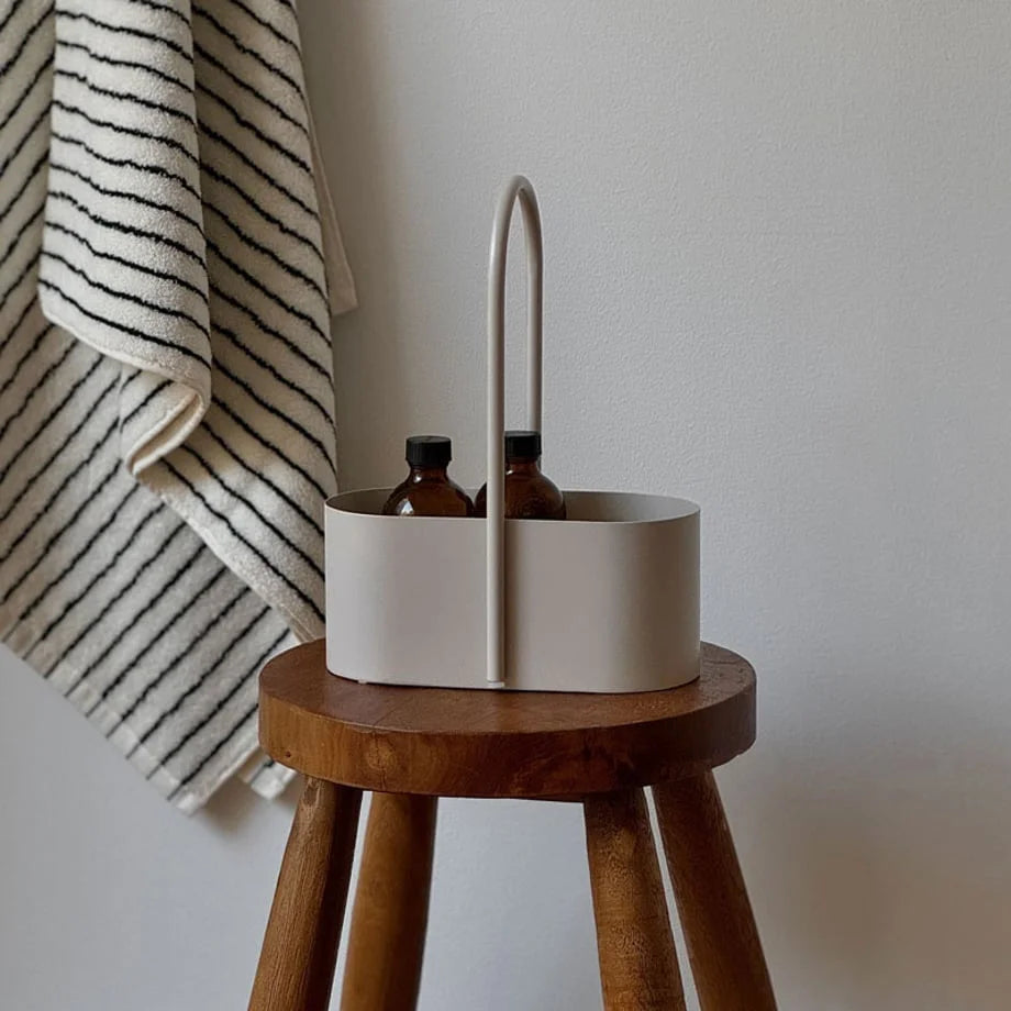 A light beige metal organizer caddy holding two amber bottles, placed on a wooden stool. - RoomDen