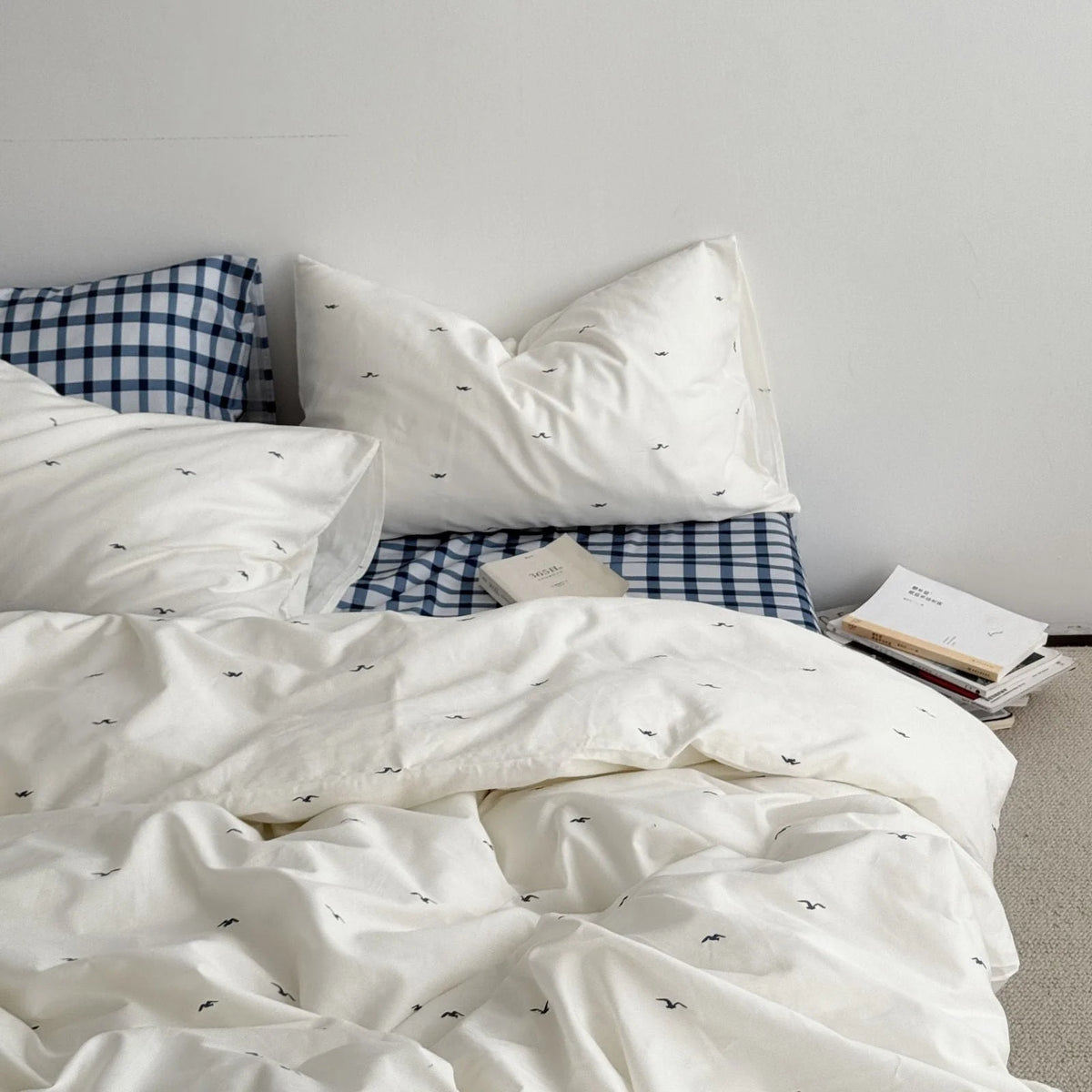 Full bedroom scene featuring the Gingham & Seagull bedding set against a white wall with books on the floor - RoomDen