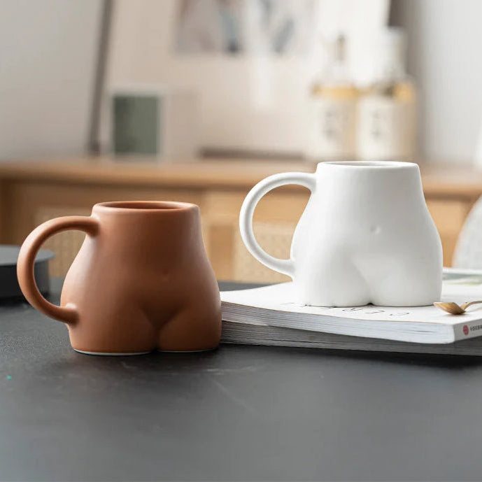 A terracotta and a white sculptural body-shaped ceramic mug on a dark table with a book. - RoomDen