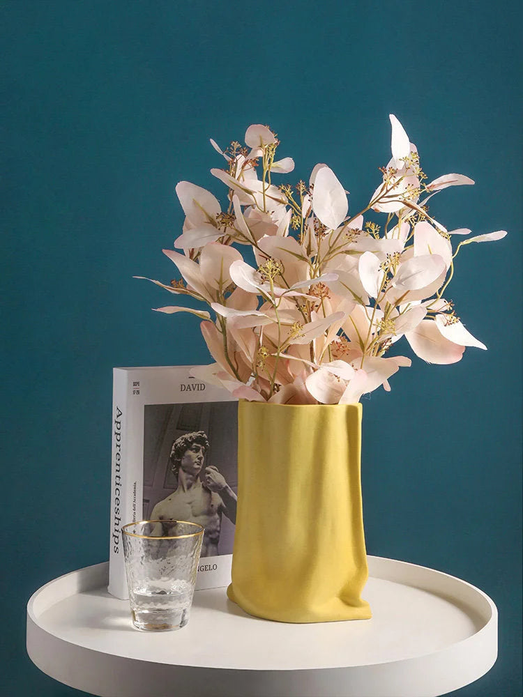 The Buttercream Yellow paper bag vase with delicate pinkish-white botanicals, styled on a white table next to a book - RoomDen.