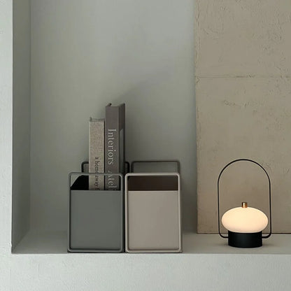 Front view of slate grey and warm beige metal organizers holding books, placed next to a modern black table lamp. - RoomDen