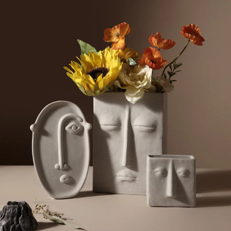Three abstract face ceramic vases in matte grey, one filled with flowers, styled on a neutral surface - RoomDen.