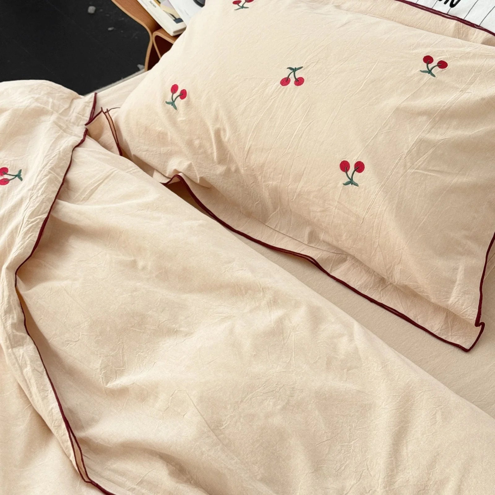 Angled shot of the creamy beige pillowcases with cherry embroidery and dark red piping.