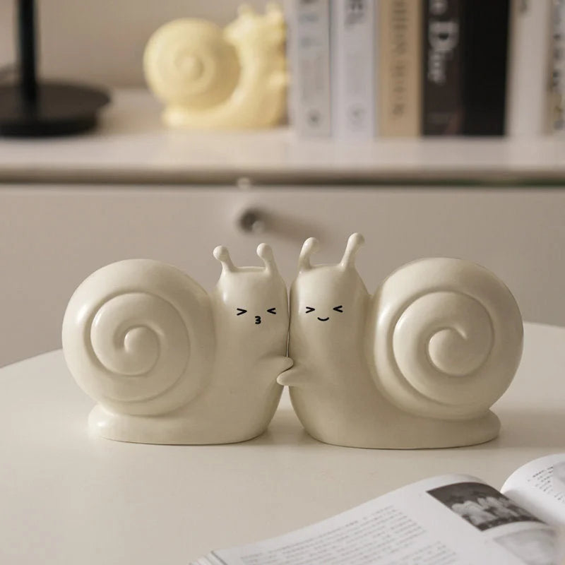A pair of creamy white snail bookends cuddling together on a white tabletop in a brightly lit room - RoomDen.