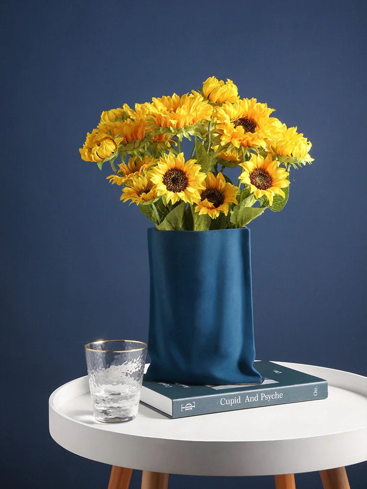 The Indigo Blue paper bag vase holding bright yellow sunflowers, styled on a white side table with a book and a glass - RoomDen.