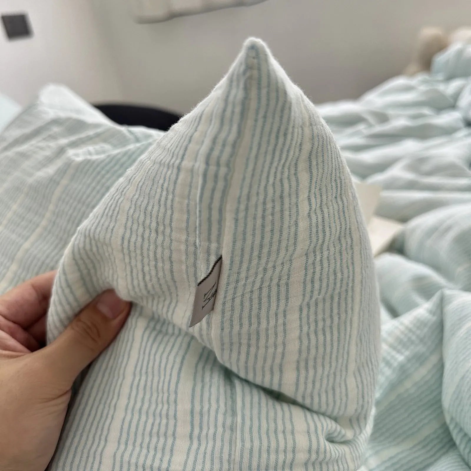 Hand holding the corner of a cyan pinstripe washed cotton pillowcase, showing the fabric's texture and tag detail.