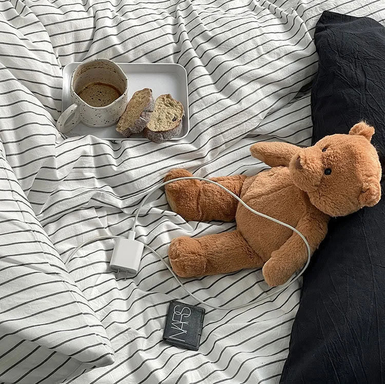 Top-down view of the pinstripe bedding with a teddy bear, a coffee cup, and a charging cable, creating a lived-in feel. - RoomDen