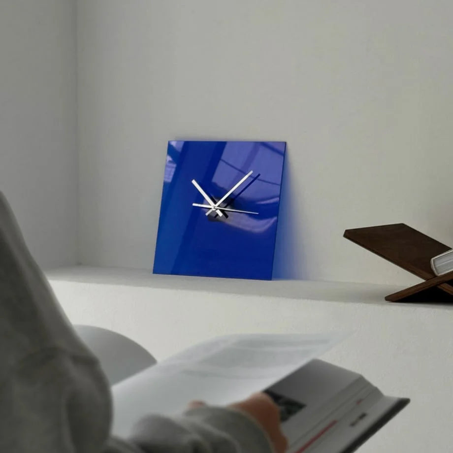 Close-up of the Cobalt Blue acrylic clock on a shelf, with a person reading in the foreground. - RoomDen