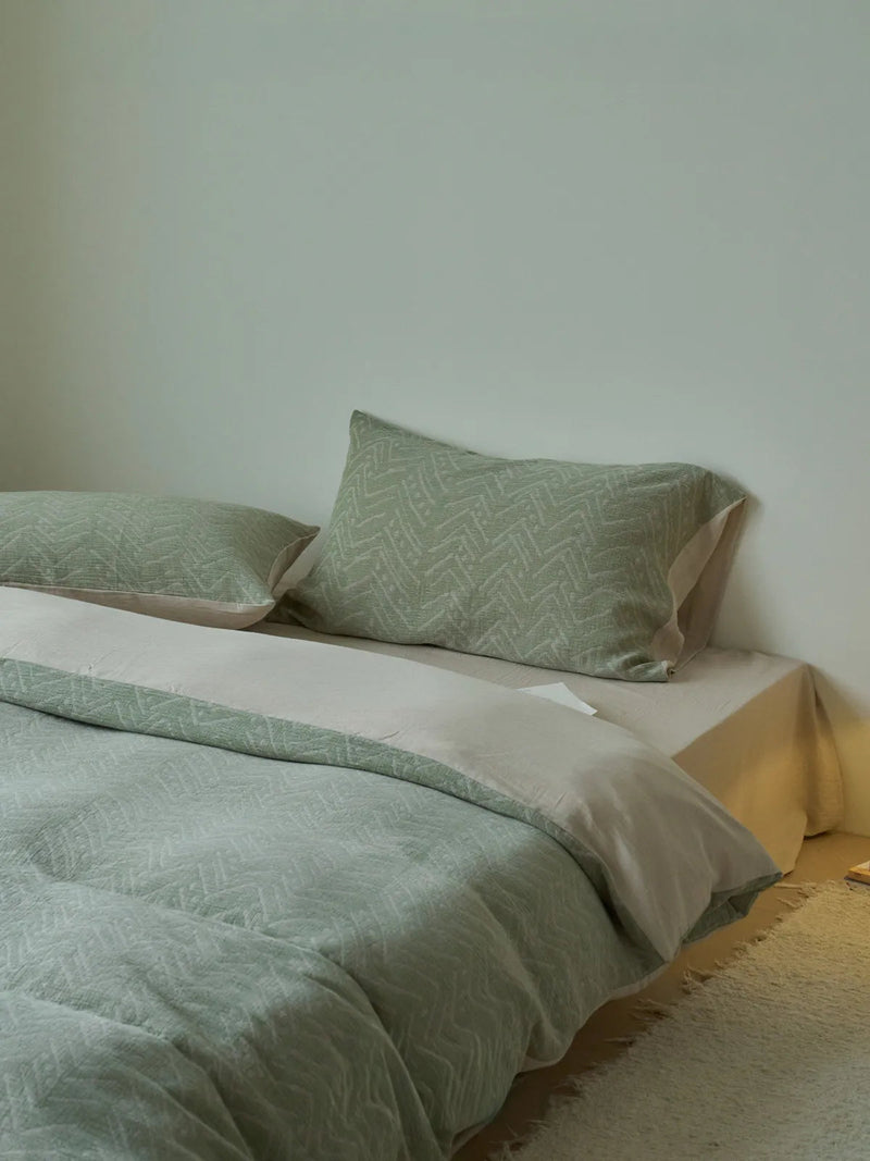 Sage green geometric bedding on a low-profile bed in a minimalist room, the beige underside of the duvet just visible. — RoomDen