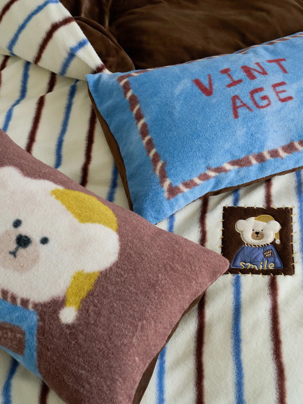 Close-up detail of the embroidered bear and 'VINTAGE AGE' text on the plush flannel fleece pillows of the bedding set. - RoomDen